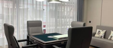 Ein modernes Zimmer mit Pooltisch, vier Stühlen, einer Couch mit Kissen und einem großen Fenster mit durchsichtigen Vorhängen.