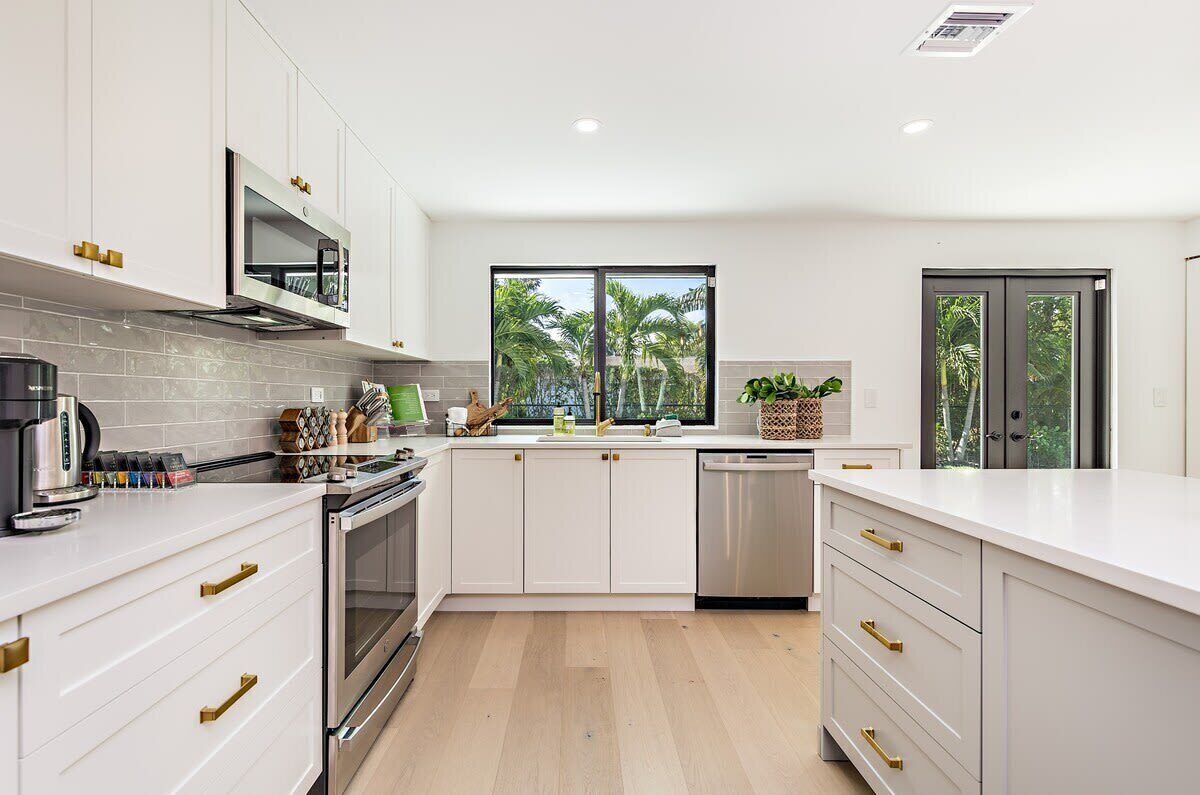 A modern kitchen with white cabinets, stainless steel appliances, and a large window with a view of palm trees.