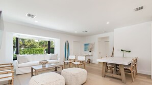 Una sala de estar moderna con un sofá blanco, una mesa de centro y una mesa de comedor con sillas. Hay un ventanal con vistas a zonas verdes.