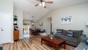 A living room with a wooden floor, a gray sofa with blue cushions, a wooden coffee table, and a ceiling fan. - AMAZING for Large Groups or Crews- 7 beds/ 4 bedrooms (Warner Robins)
