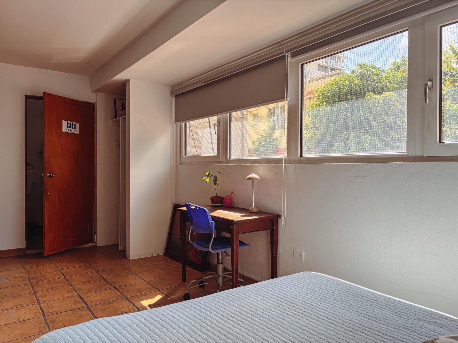 A room with a bed, a desk with a chair, a window with a view of greenery, and a door labeled "BANOS".