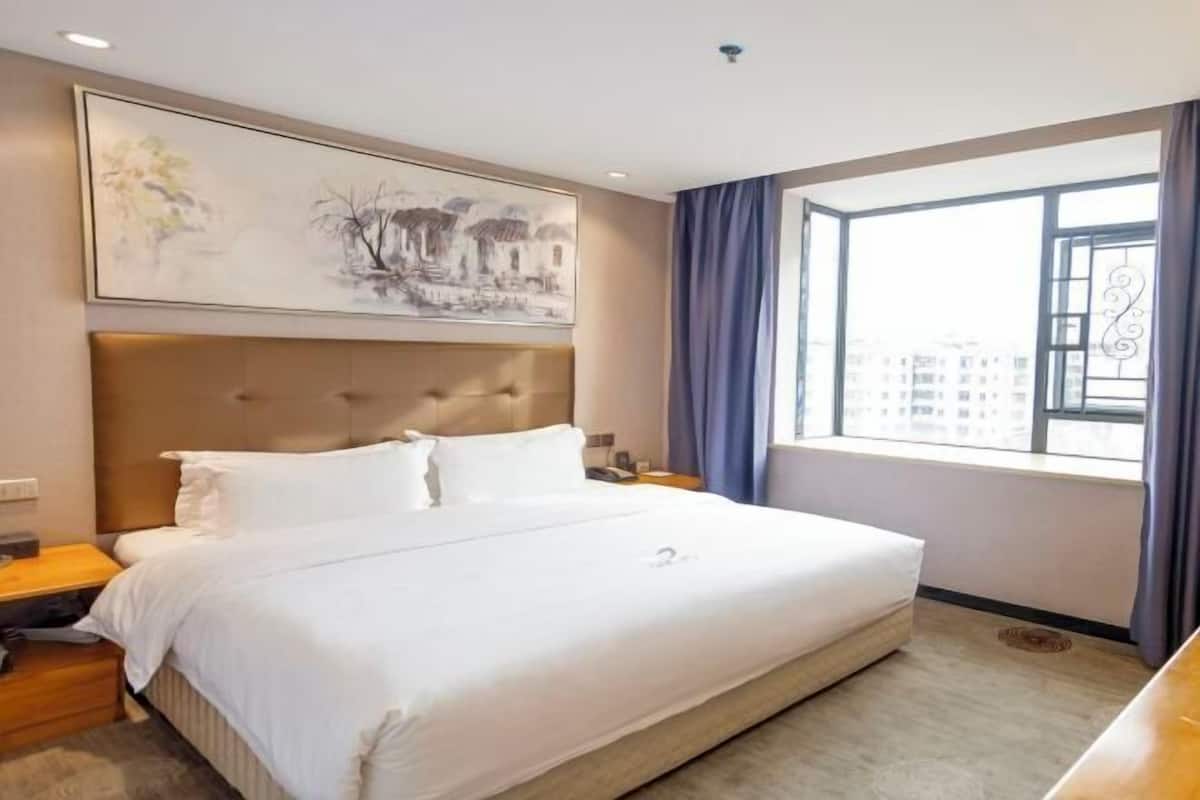 A hotel room with a large bed, a headboard, a nightstand, a painting on the wall, a window with curtains, and a view of buildings outside.