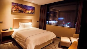 Habitación de hotel con una cama, una mesita de noche, una silla, un cuadro y una ventana con vistas a la ciudad.