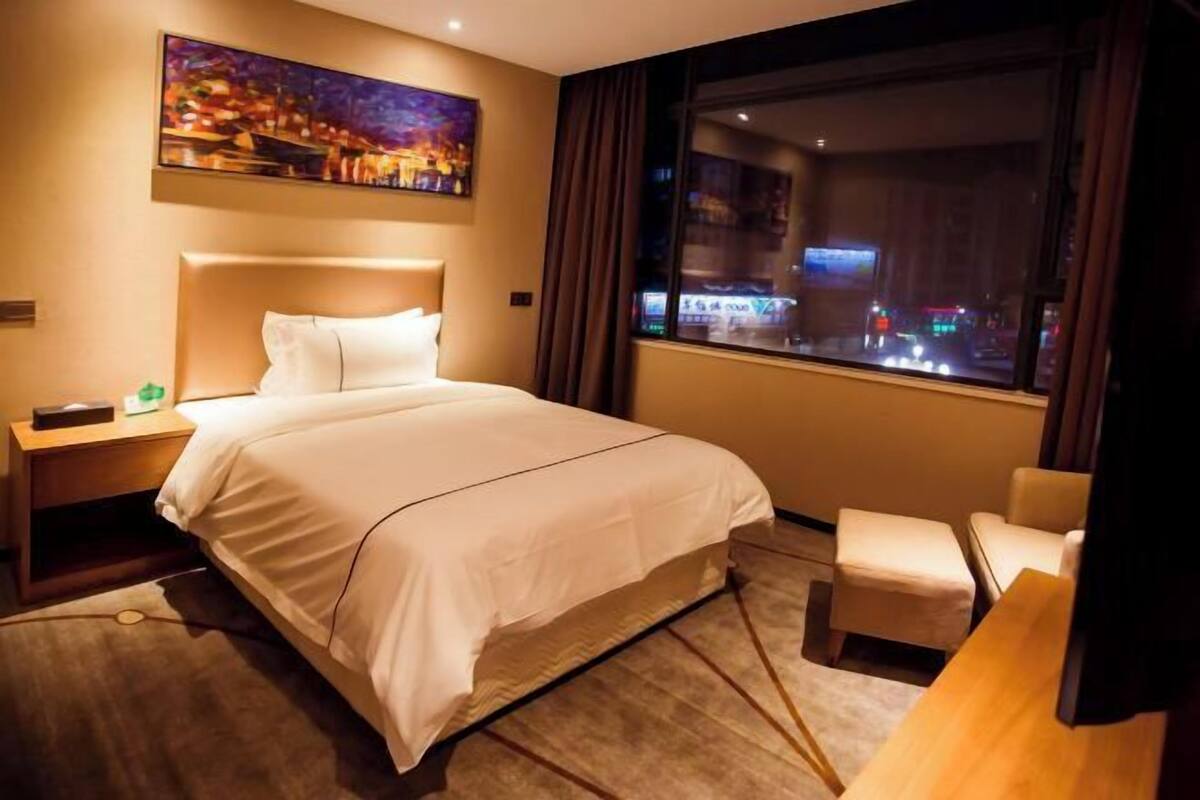 A hotel room with a bed, a nightstand, a chair, a painting, and a window with a city view.