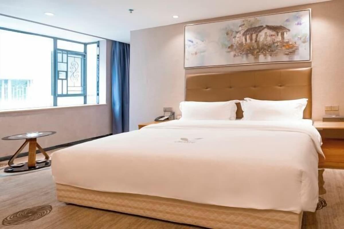 Una habitación de hotel con una cama grande, una ventana con cortinas, un cuadro en la pared y una mesita redonda con una lámpara.