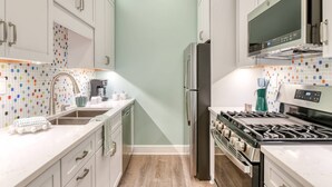 A narrow kitchen with white cabinets, a stainless steel refrigerator, a microwave, and a gas stove. - Brand New! Heart of Historic St. Augustine, Walk Everywhere, 4 Miles to Beach, Parking Spot, Balcony (St. Augustine)