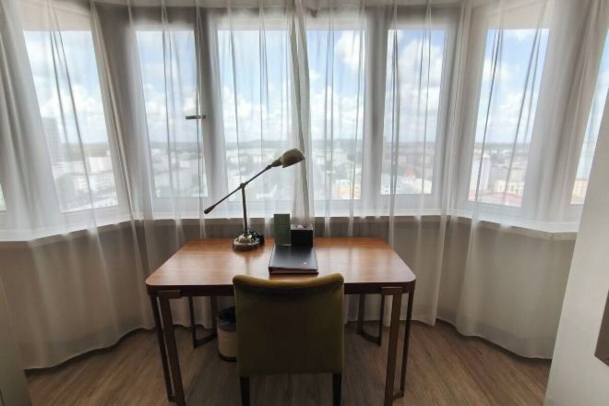 Ein Zimmer mit einem Holztisch, einem Stuhl, einer Lampe und Blick durch leichte Vorhänge.