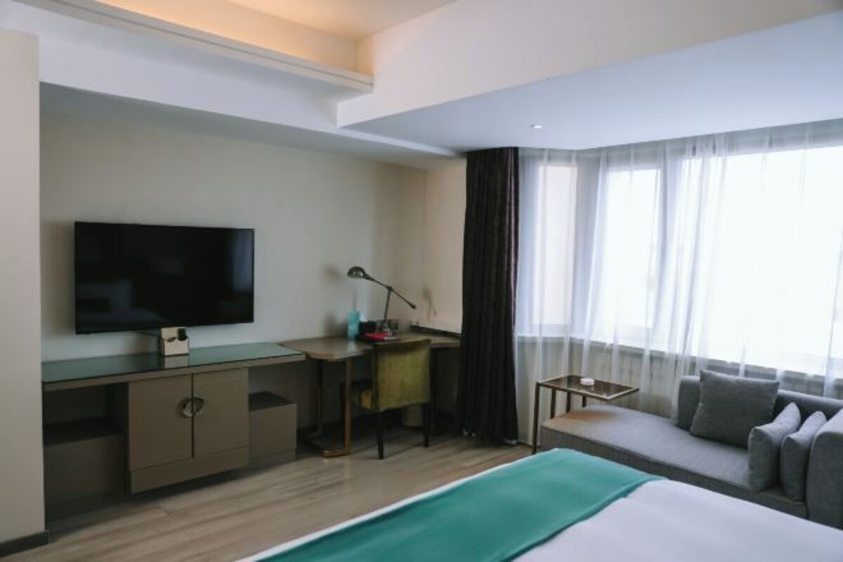 A hotel room with a bed, a desk with a TV, a chair, a sofa, and a window with curtains.