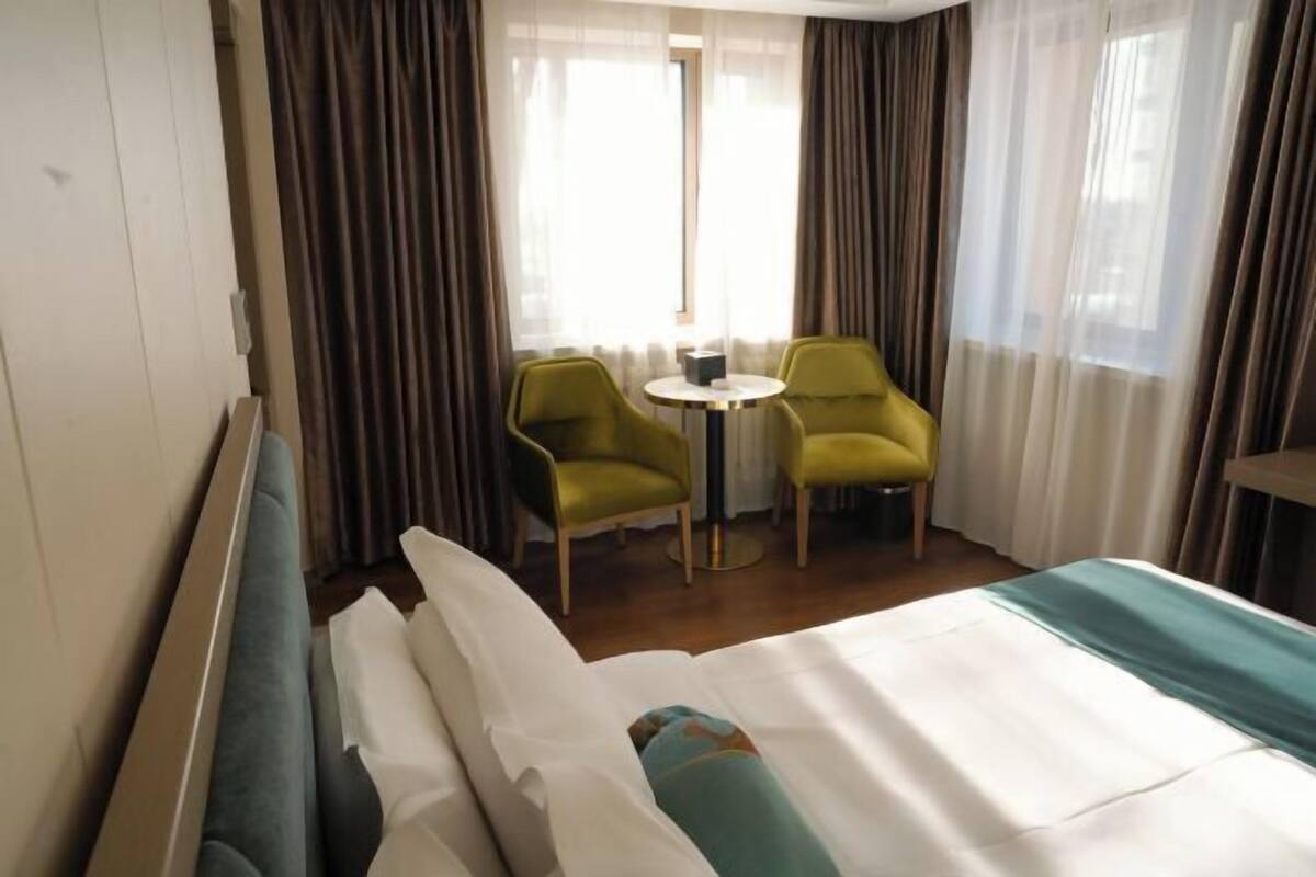 Hotellihuone, jossa on sänky, kaksi vihreää nojatuolia, pieni pyöreä pöytä ja ikkuna, jossa on verhot.