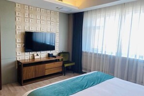 A hotel room with a large flat-screen TV mounted on a textured wall, a wooden cabinet with drawers, a green chair, a bed with a teal bedspread, and a large window with sheer curtains. - Runsha International Hotel (Hegang)