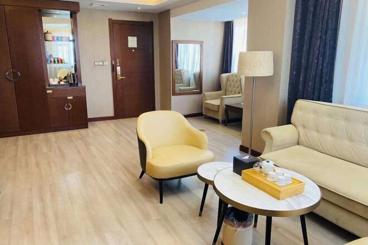 A hotel room with a wooden wardrobe, a yellow armchair, a beige sofa, a small round table with a tray of items, and a floor lamp.