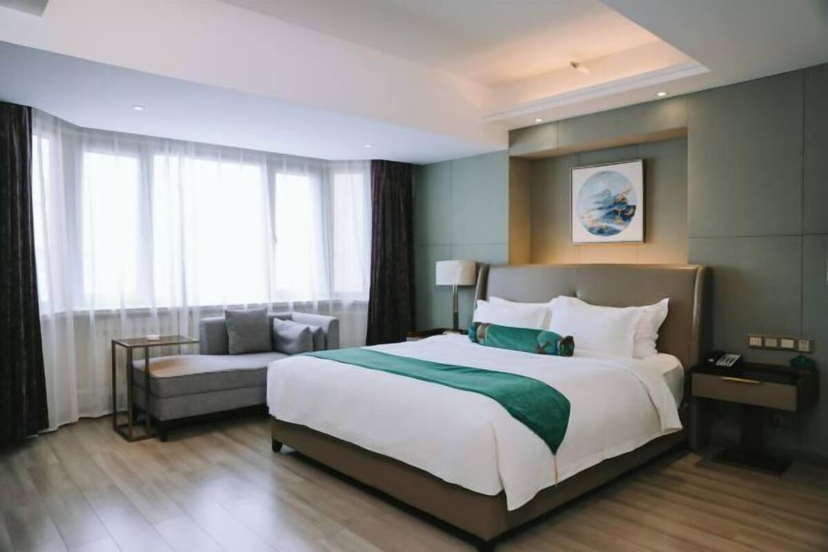 A hotel room with a large bed, a bedside table, a lamp, a painting on the wall, and a view of the outside through the window.