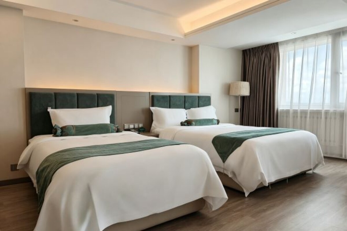 A hotel room with two beds, a large window with curtains, and a lamp on the nightstand.