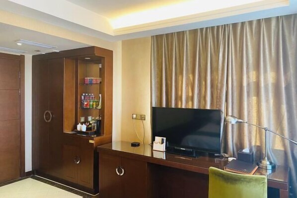 A hotel room with a wooden wardrobe, a flat-screen TV, a desk with a lamp, and a large window with curtains. - Runsha International Hotel (Hegang)