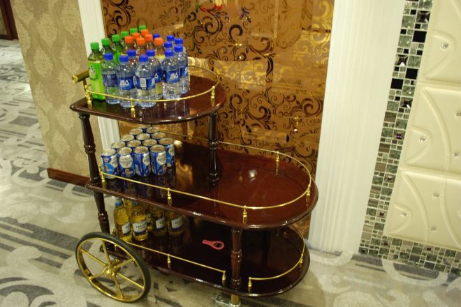 A three-tiered cart with bottles and cans, positioned in a room with patterned wallpaper and a tiled floor.