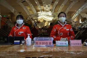 Two staff members in red uniforms standing behind a reception desk with a nameplate, a plastic container, and a bottle of hand sanitizer.