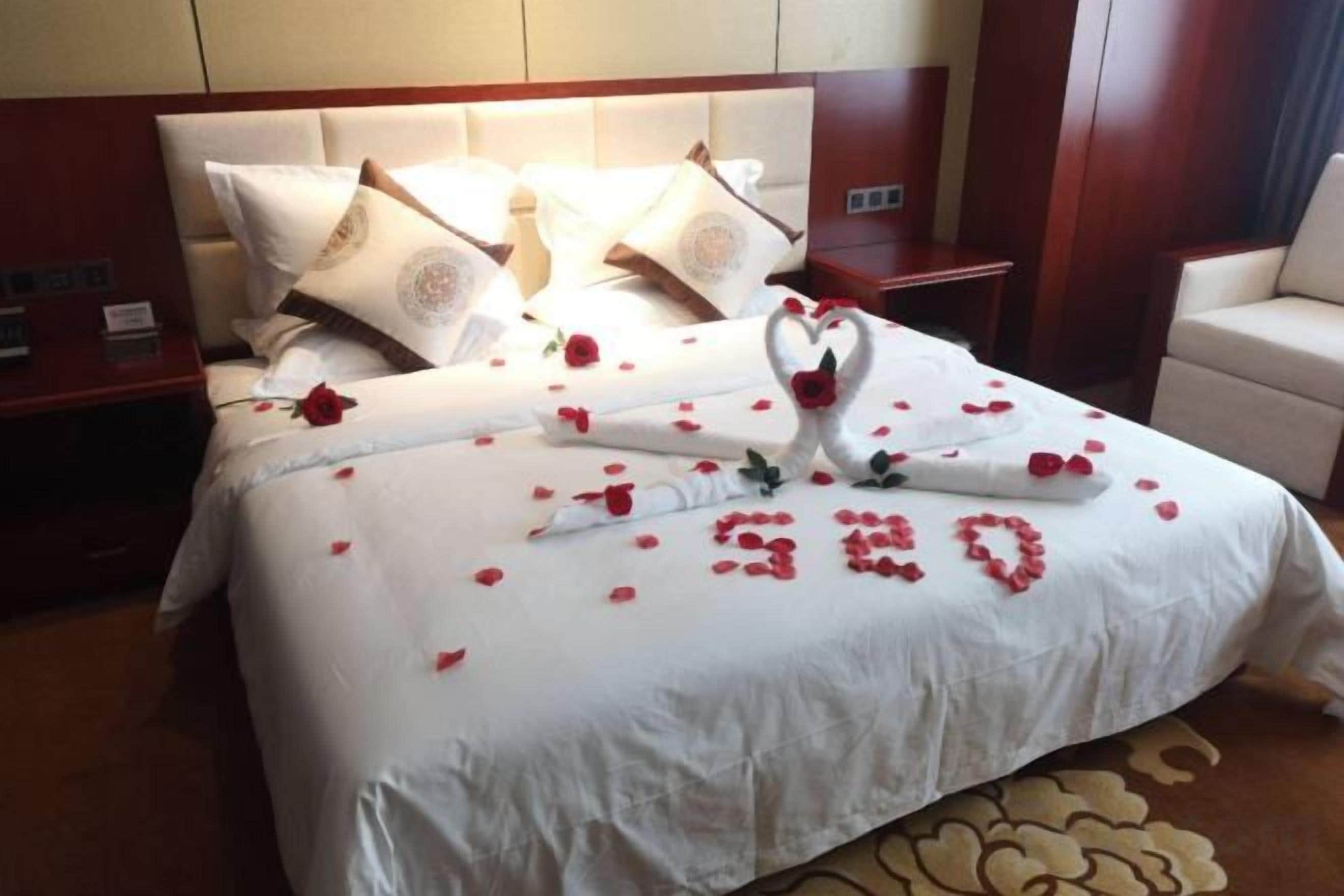 A neatly made bed with a white bedspread, pillows, and a decorative arrangement of rose petals and a heart-shaped swan.