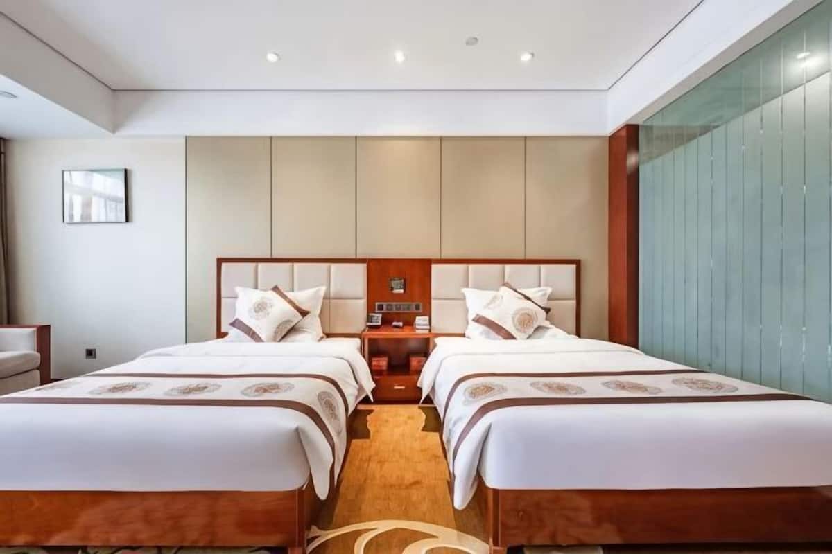 Quarto de hotel com duas camas, uma mesa de cabeceira, um relógio fixado na parede e uma janela grande.