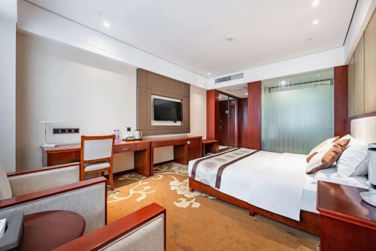 A hotel room with a bed, desk, chair, TV, and a shower area.