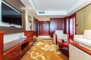 Un vestíbulo de hotel con un televisor de pantalla plana montado en la pared, un mostrador de recepción, una zona de estar con un sofá y sillones, y una alfombra estampada.