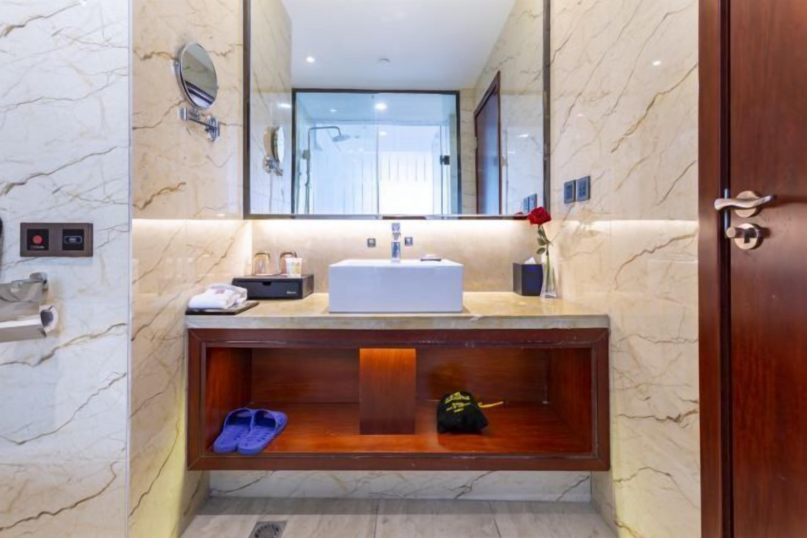 A modern bathroom with a marble vanity, a large mirror, and a wooden shelf with slippers.