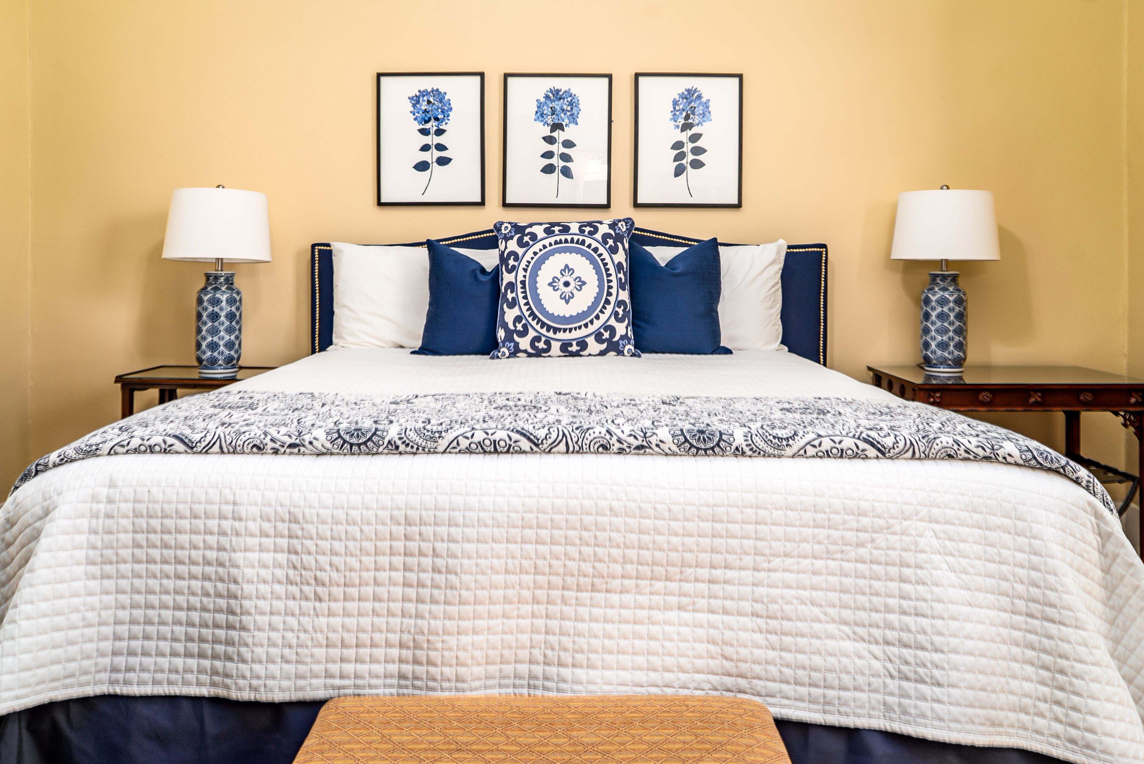 Un lit bien fait, recouvert d’une couvre-lit à motifs, avec deux lampes assorties et trois œuvres d’art florales encadrées accrochées au mur.