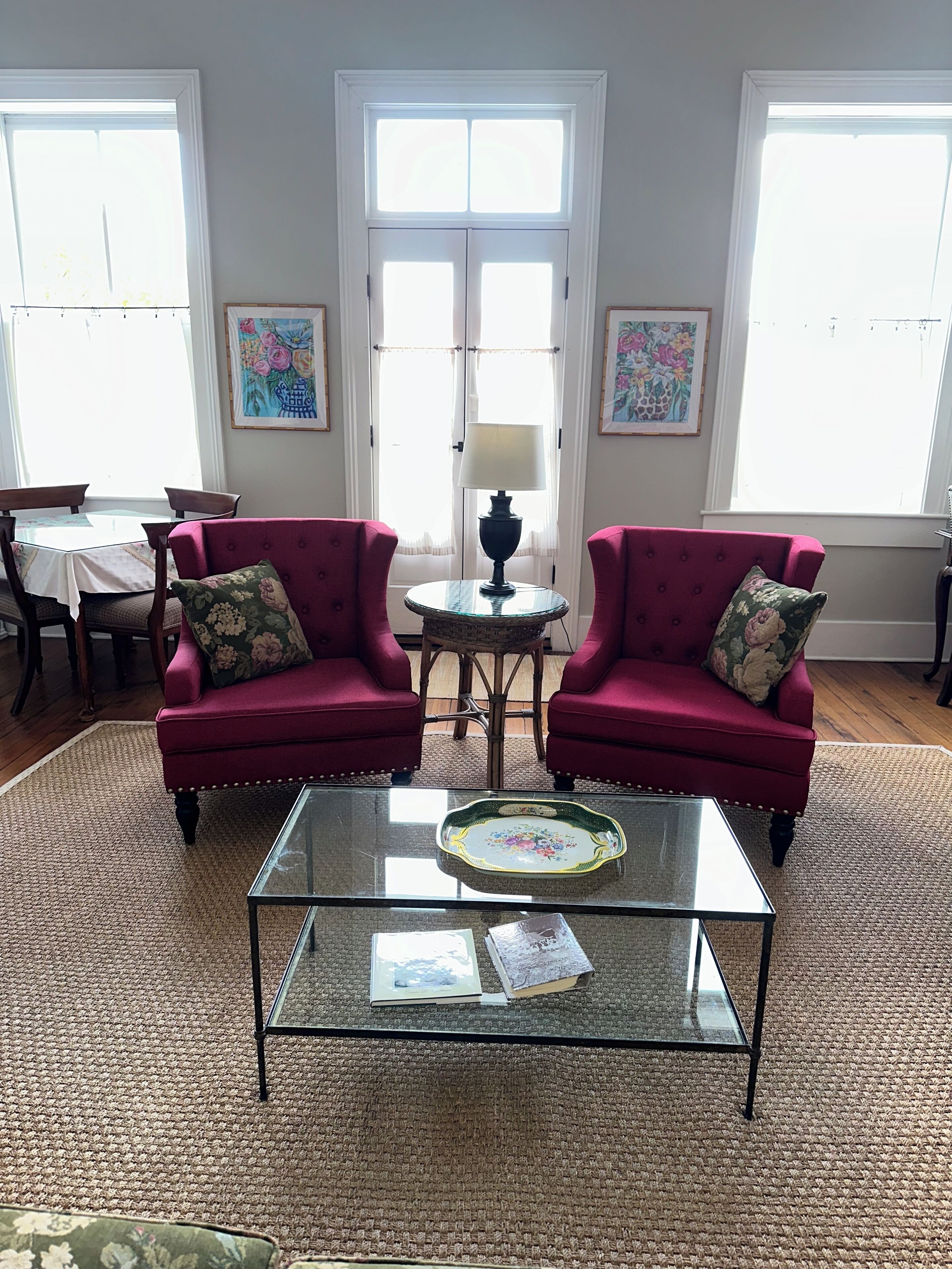 Un salon avec deux fauteuils rouges, une table basse en verre, une petite table ronde avec une lampe, et une table à manger avec des chaises.