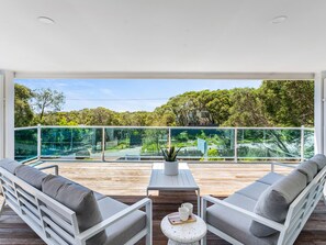 A balcony with a wooden deck, two grey sofas, a white table, and a glass railing overlooking a forested area. - Rye Baby by Ready Set Stay- Main & Guest House (Rye)