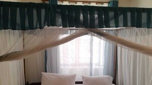 Un lit simple avec une literie blanche et deux oreillers, un ventilateur de plafond et des rideaux légers.