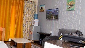 Un salon comprenant un canapé, une table basse, un distributeur d’eau, un réfrigérateur et un téléviseur à écran plat fixé au mur.