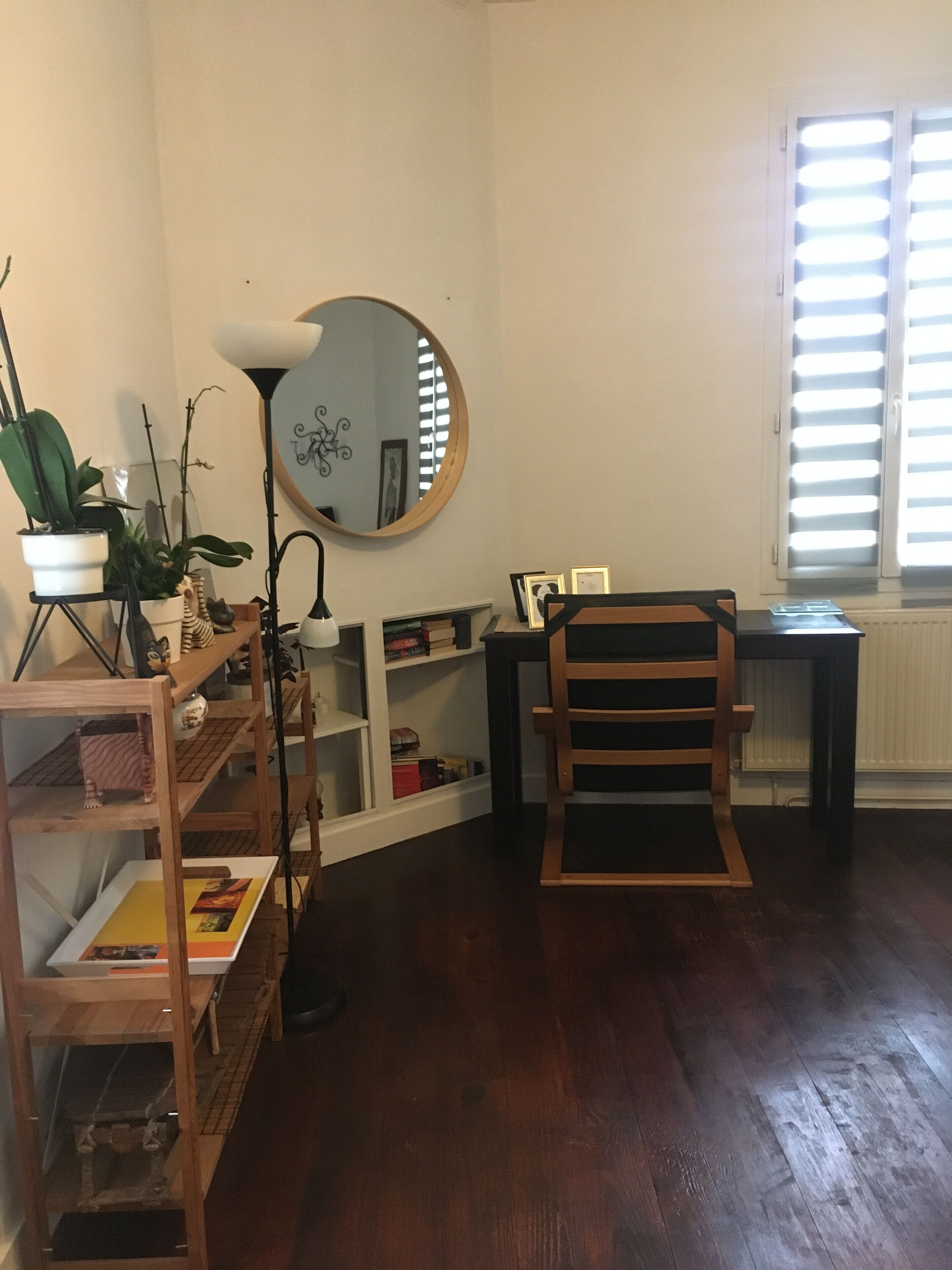 A room with a wooden floor, a desk with a chair, a mirror, shelves with books and decorative items, and a window with blinds.
