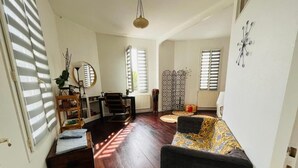 Un salon avec un canapé foncé, un plancher en bois, une petite table et une chaise. On y trouve une fenêtre avec des stores, un élément décoratif mural et un luminaire suspendu.