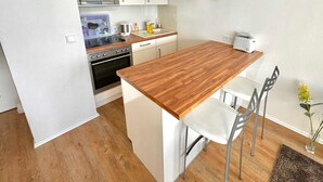 Une cuisine moderne avec des armoires blanches, un plan de travail en bois, un four encastré, un four à micro-ondes et un grille-pain. On y trouve deux chaises blanches et une petite table.