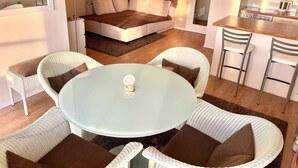 Un salon moderne avec une table ronde en verre, des fauteuils en osier blancs, un canapé avec des coussins marron et blancs, et un coin cuisine équipé d’une cuisinière et de chaises.