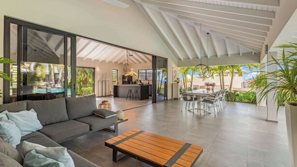 Un vaste espace de vie comprenant un canapé, une table basse et une table à manger avec des chaises. Au fond, on trouve un coin cuisine avec un bar et des tabourets.