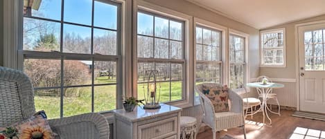 A sunlit room with large windows, wicker furniture, a small table, and a white door.