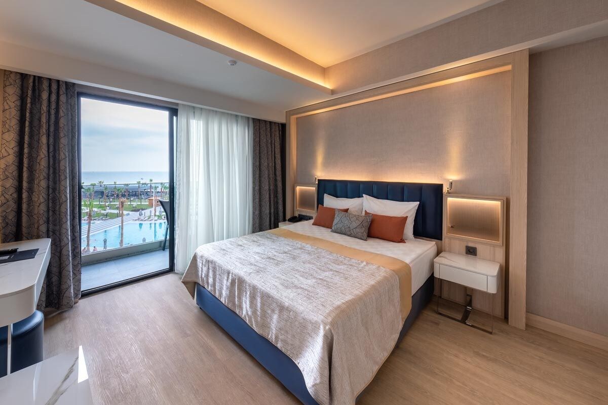 A hotel room with a large bed, bedside tables, a desk, and a view of a pool and cityscape.