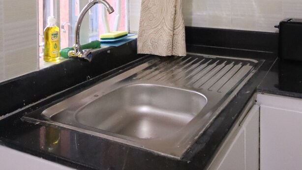 A kitchen with a stainless steel sink, a window with a curtain, and white cabinets. - Stay.Plus Ebony Mara House (Narok)