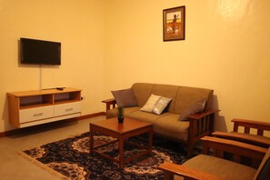 A living room with a sofa, two wooden side tables, a coffee table, a TV on a stand, and a framed picture on the wall. - Stay.Plus Ebony Mara House (Narok)