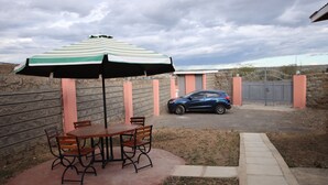 Une terrasse avec une table et des chaises, un parasol à rayures et une voiture garée au fond.