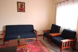 A living room with a dark blue sofa, two wooden armchairs, a small wooden coffee table with a plant, and a red patterned rug. - Stay.Plus Ebony Mara House (Narok)