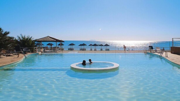 A clear swimming pool with two people in the center, surrounded by lounge chairs and umbrellas, with a view of the sea and a small pavilion in the background.