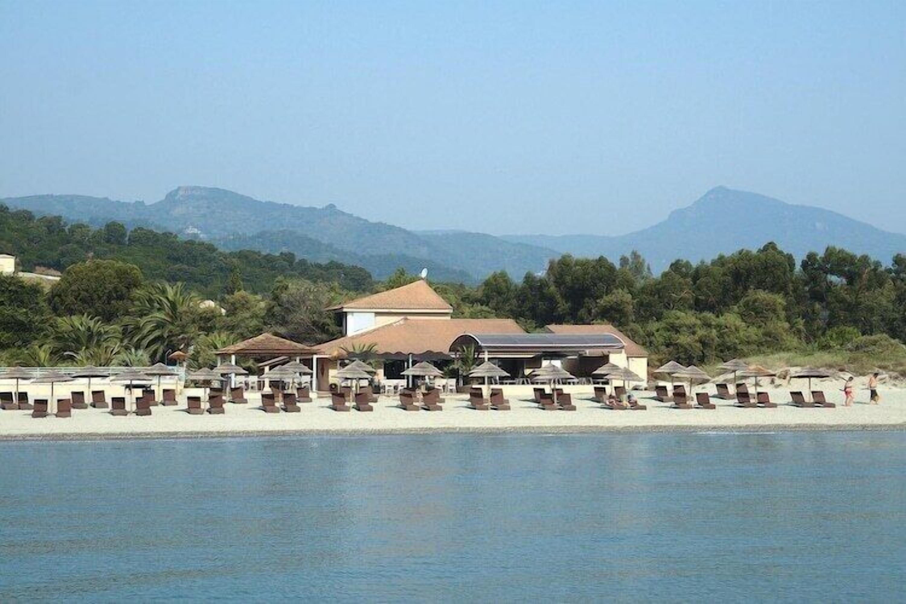 A beachside resort with sun loungers and umbrellas, a building with a red roof, and mountains in the background.