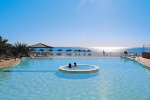 Een helder zwembad met een rond bubbelbad in het midden, omringd door ligbedden en parasols, met uitzicht op de zee op de achtergrond.