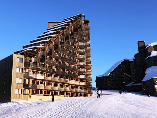 A multi-story building with balconies, a ski slope in the background, and snow on the ground.