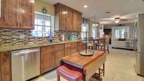 A kitchen with wooden cabinets, a stainless steel dishwasher, a wooden dining table with a red bench, and a tiled backsplash. - Riverfront Getaway w/ Hot Tub & Game Room! (New Port Richey)