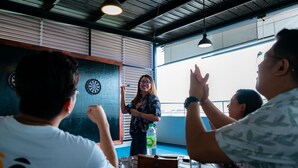 A group of people at a table with a dartboard, a water bottle, and a clock on the wall. - JPOD Capsule Hotel (Kota Kinabalu)