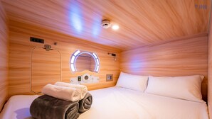 A hotel room with a wooden ceiling, a bed with white linens, a towel and robe on the bed, a wall-mounted mirror, and a ceiling light fixture. - JPOD Capsule Hotel (Kota Kinabalu)