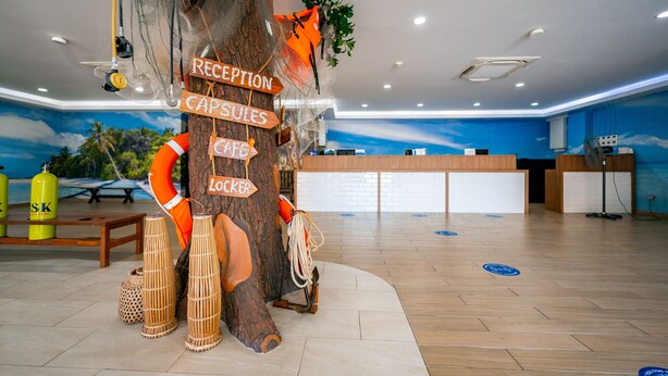 A reception desk area with a wooden tree trunk centerpiece, signs for reception, capsules, cafe, and locker. - JPOD Capsule Hotel (Kota Kinabalu)