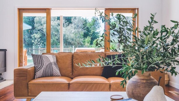 Ein Wohnzimmer mit einem braunen Ledersofa, einem weißen Couchtisch und einem großen Fenster mit Blick auf Grünfläche.